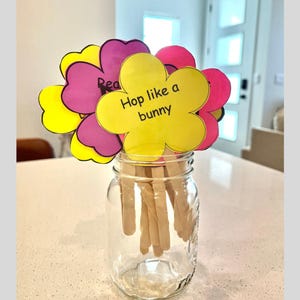May include: A clear glass jar filled with wooden sticks, holding colorful paper flowers. The flowers are yellow, pink, and purple, with the words "Hop like a bunny" and "Read" written on them. The jar sits on a white countertop.