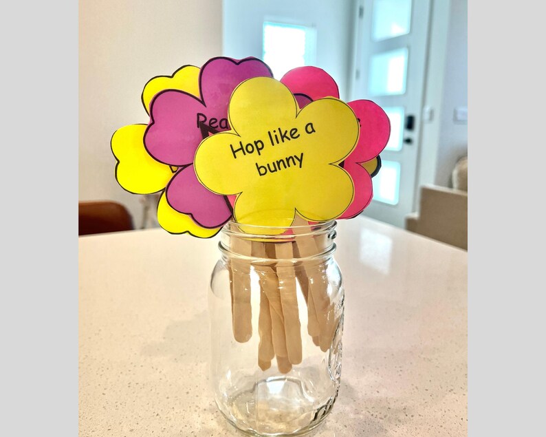 May include: A clear glass jar filled with wooden sticks, holding colorful paper flowers. The flowers are yellow, pink, and purple, with the words "Hop like a bunny" and "Read" written on them. The jar sits on a white countertop.
