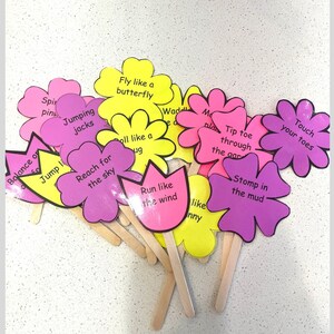 May include: A collection of colorful flower-shaped cards on wooden sticks. Each card features a different action prompt, such as "Fly like a butterfly" and "Touch your toes", in black text. The flowers are pink, yellow, and purple.