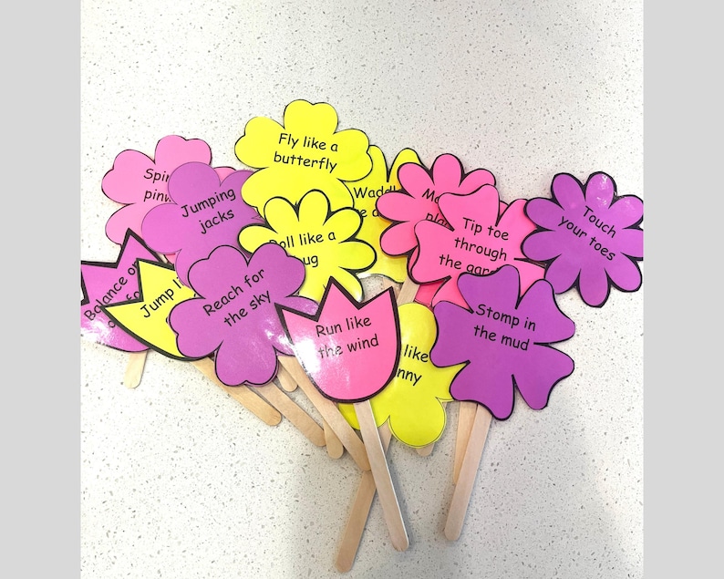 May include: A collection of colorful flower-shaped cards on wooden sticks. Each card features a different action prompt, such as "Fly like a butterfly" and "Touch your toes", in black text. The flowers are pink, yellow, and purple.