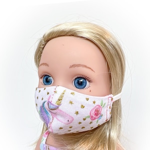 May include: A white doll wearing a pink, white, and gold unicorn face mask with white polka dots. The mask has a pink rose and green leaves on one side.