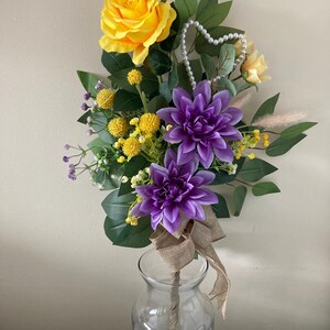 May include: A bouquet of artificial flowers in a clear glass vase. The bouquet includes a yellow rose, purple dahlias, yellow button flowers, and green leaves. A pearl heart-shaped garland is wrapped around the stems.