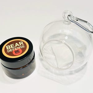 May include: A small, round, brown jar with a black lid and a label that reads "BEAR DEFENSE BE ARMED". The jar is next to a clear glass orb with a silver clip.