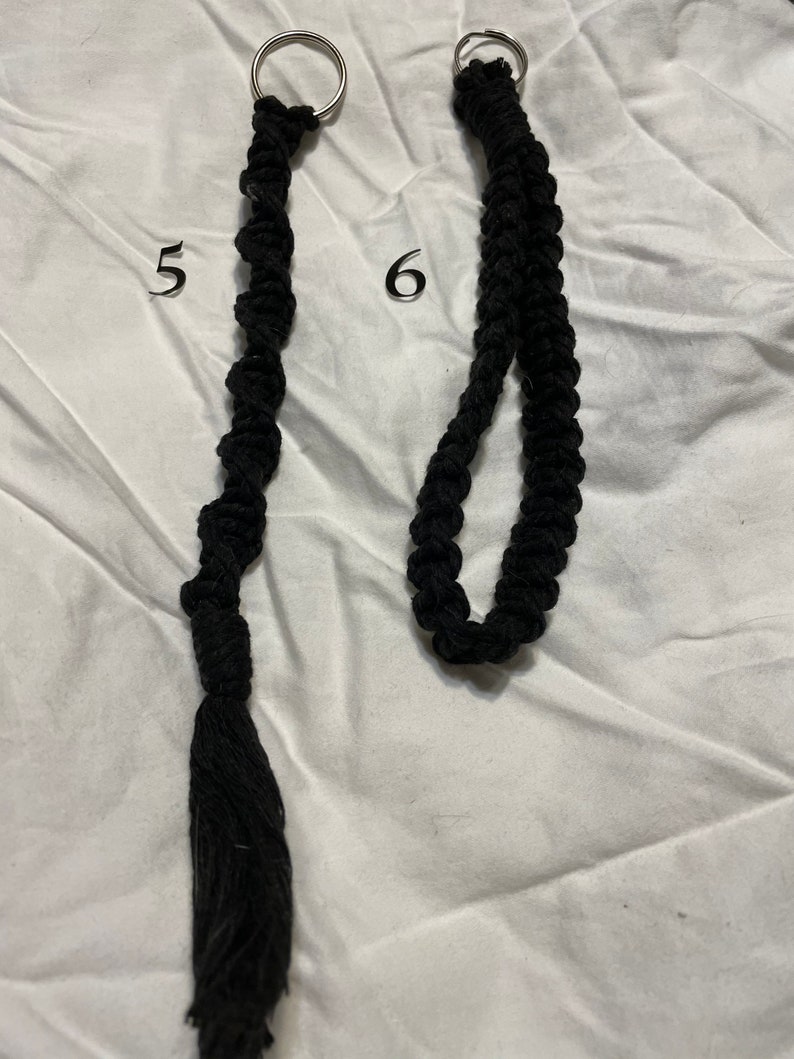 May include: Two black macrame keychains with silver key rings. The keychain on the left has a tassel at the end. The keychain on the right has a loop at the end.