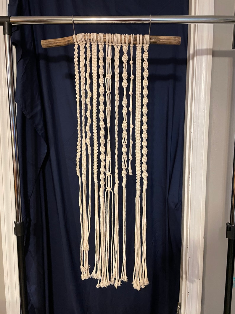 May include: A white macrame wall hanging with a variety of knots and patterns. The hanging is attached to a wooden dowel rod.