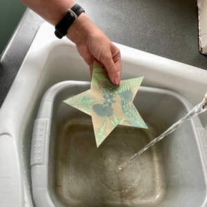 May include: A hand holds a star-shaped paper craft with a floral pattern over a grey basin filled with water. A stream of water flows from a tap into the basin.
