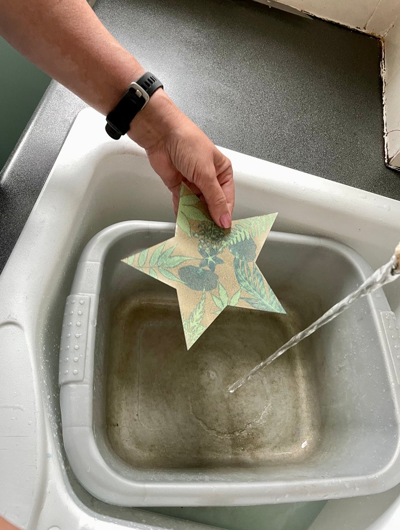 May include: A hand holds a star-shaped paper craft with a floral pattern over a grey basin filled with water. A stream of water flows from a tap into the basin.