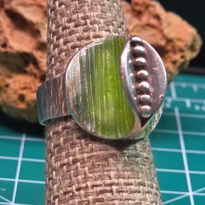 Puede incluir: Un anillo de plata con una cara circular que presenta un panel central de color verde lima texturizado y una fila vertical de pequeñas esferas de plata en el lado derecho. La banda del anillo tiene una textura acanalada vertical.