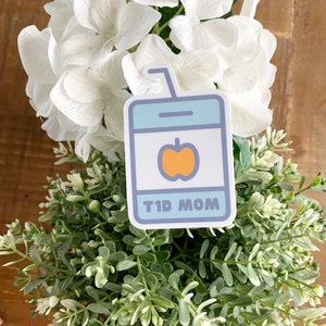 Pode incluir: Um autocolante com um design de caixa de sumo e um gr&aacute;fico de ma&ccedil;&atilde;. A caixa de sumo &eacute; azul claro com um centro branco e uma ma&ccedil;&atilde; laranja. O texto "T1D MOM" &eacute; exibido na parte inferior do autocolante. O autocolante &eacute; colocado sobre uma cama de folhagem verde e branca.