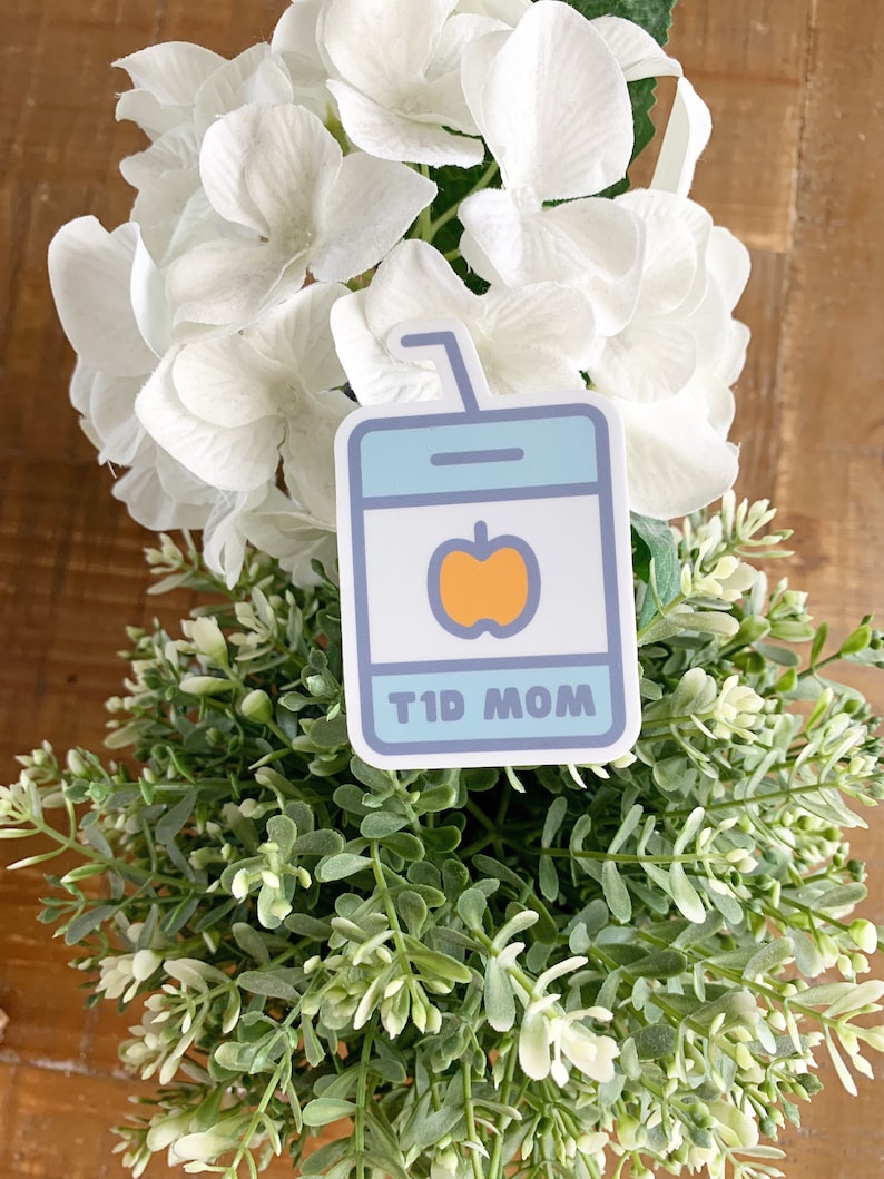 Pode incluir: Um autocolante com um design de caixa de sumo e um gr&aacute;fico de ma&ccedil;&atilde;. A caixa de sumo &eacute; azul claro com um centro branco e uma ma&ccedil;&atilde; laranja. O texto "T1D MOM" &eacute; exibido na parte inferior do autocolante. O autocolante &eacute; colocado sobre uma cama de folhagem verde e branca.