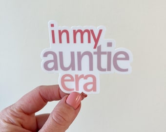 May include: A white sticker with the text "in my auntie era" in pink and red lettering.
