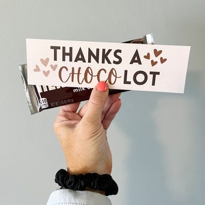 May include: A white card with the text "Thanks a Choco Lot" in brown lettering and pink hearts. The card is held in front of a chocolate bar.