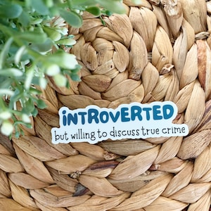 May include: A sticker with the text "INTROVERTED but willing to discuss true crime" in white and blue lettering. The sticker is placed on a woven, light brown surface, with green foliage in the upper left corner.