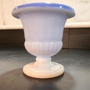 May include: A light blue and white urn-shaped vase with a wide, rounded top and a pedestal base. The vase features a detailed, embossed floral design on the body. The base has a peach-colored rim.