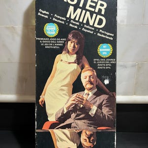 May include: A rectangular board game box for Original Master Mind. The black box features a photograph of two people, text in multiple languages, and two yellow award seals. The bottom includes the text A GAME OF CUNNING AND LOGIC FOR TWO PLAYERS BREAK THE HIDDEN CODE and four colored circles.