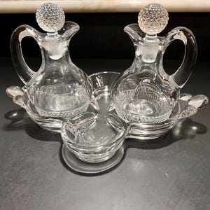 May include: A clear glass cruet set featuring two small pitchers with textured spherical stoppers and curved handles. The pitchers sit within a matching three-sectioned glass tray that includes a central bowl, all with a diamond-patterned base.