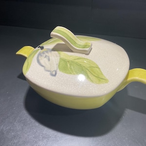 May include: A ceramic apple-shaped serving dish with a lid. The dish is primarily yellow with a cream-colored lid decorated with green leaves and a blue fruit design. The lid has a green handle.