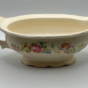 May include: A white ceramic gravy boat with a floral design in shades of pink, yellow, and blue. The boat has a silver rim and a scalloped edge.