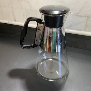May include: A clear glass carafe with a black lid and handle, and a silver-colored band around the middle. The carafe has a spout for pouring. The handle is attached to the side with silver rivets. The carafe is sitting on a gray surface.