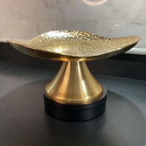 May include: A gold-colored decorative bowl with a textured surface, resting on a pedestal base. The bowl has a wide, shallow design, and the base is a contrasting black color. The bowl is likely made of metal.