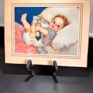 May include: Vintage print of a baby in a diaper and blue outfit, holding a bottle. The baby is lying on a pink pillow with a blue background. The print is framed in a beige border and displayed on a black stand.