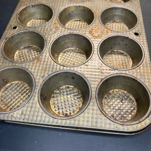 May include: A vintage metal muffin tin with 12 individual cups. The tin is worn and has a speckled brown patina.