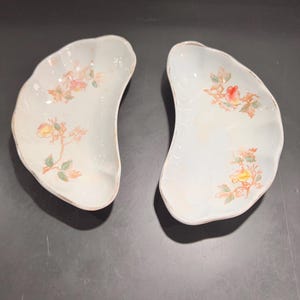 May include: Two white ceramic half-moon shaped serving dishes with floral designs in shades of yellow, orange, and green.