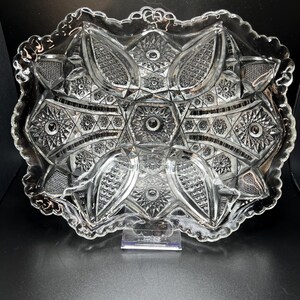 May include: A rectangular clear glass serving tray featuring an intricate pressed pattern of starbursts, cross-hatching, and geometric shapes. The tray has a scalloped, decorative edge and a central hexagonal motif with a starburst design.