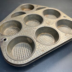 May include: A vintage metal muffin tin with six individual molds. The tin is gray and has a textured surface. Each mold has a raised, diamond-shaped pattern.