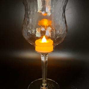 May include: A clear glass candle holder with a textured surface and a long stem. An orange LED candle is inside the holder, which is lit. The candle holder is on a black background.