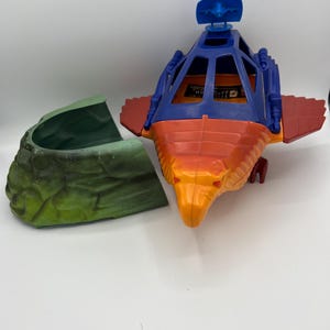 May include: A vintage He-Man action figure spaceship with a green rocky base. The spaceship is orange, blue, and red with a cockpit and a star on top.
