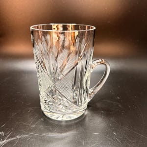 May include: Clear crystal glass mug with a handle. The mug features a detailed pattern of etched lines and geometric shapes. The glass has a slightly tapered shape and a rounded handle. The mug is designed for beverages.