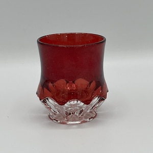 May include: A small, ruby red glass with a clear, patterned base. The upper portion of the glass is a deep red, while the base features a detailed, cut-glass design. The glass is likely a decorative item or a small drinking vessel.