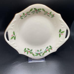 May include: Cream-colored serving dish with a gold rim and holly leaf and berry pattern. The dish has a square shape with rounded corners and handles on either side. The holly design features green leaves and red berries.