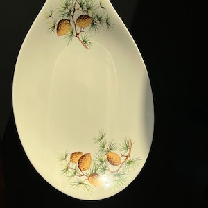 May include: A white oval serving dish with a green and brown pine cone design. The dish has a slightly curved edge.