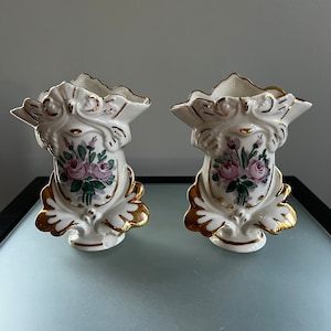 May include: A pair of white porcelain vases with gold accents and floral designs. The vases have a scalloped top and a base with a decorative design. The flowers are pink and green.