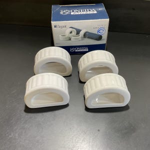 May include: Four white ceramic napkin rings with a ridged design, arranged in front of a box. The box features the words "ONEIDA Kitchen" and "Elegant" and shows a picture of the napkin rings. The napkin rings are a kitchen accessory.