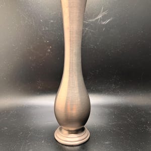 May include: A slender, metallic vase with a brushed finish. The vase has a flared top, a rounded midsection, and a circular base. The overall shape is elongated, with a smooth, polished appearance. The vase is a muted silver color.