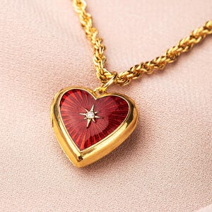 Könnte beinhalten: Eine goldene herzförmige Anhängerkette mit rotem Emaille-Mittelteil und kleinem Diamantakzent. Der Anhänger hängt an einer dicken, strukturierten Goldkette. Das Herzdesign hat ein Sonnenstrahlenmuster.
