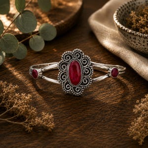 Puede incluir: Un brazalete de plata con una gran piedra preciosa roja ovalada en el centro rodeada de intrincados adornos metálicos. La banda del brazalete se divide en dos brazos, cada uno con una pequeña piedra preciosa roja redonda a los lados.