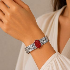 Puede incluir: Una pulsera tipo brazalete de plata con una piedra roja ovalada en el centro que presenta un patrón floral en relieve. La banda está hecha de plata oxidada con intrincados grabados geométricos y florales a lo largo de toda la superficie.