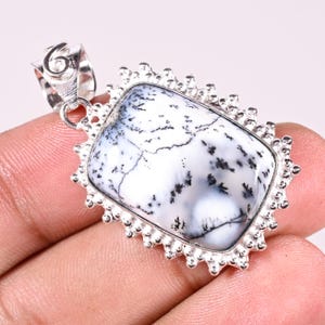 May include: A rectangular pendant featuring a white dendritic agate stone with black fern-like inclusions. The stone is set in a decorative silver frame with a scalloped edge and a textured bail at the top for attaching to a chain or cord.