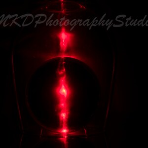May include: A red light shines through a clear glass bottle, creating a glowing red orb inside. The light is reflected on the bottom of the bottle.