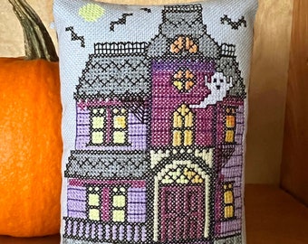 Puede incluir: Una almohada bordada de color morado y gris con un diseño de casa encantada. La casa tiene una puerta, ventanas y un fantasma. Hay murciélagos y una luna en el fondo. La almohada está decorada con dos calabazas.