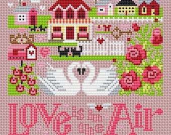 Puede incluir: Patrón de punto de cruz con una encantadora escena de pueblo con casas, cisnes y rosas. El texto "Love is in the Air" está bordado en rosa debajo de la imagen.