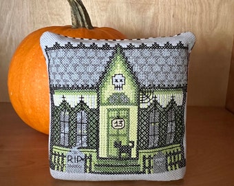 Puede incluir: Una almohada bordada en gris y verde con un diseño de casa espeluznante. La casa tiene una calavera en el techo, una puerta con el número 13 y un gato negro sentado en el porche. Las palabras "RIP" están bordadas en el porche.