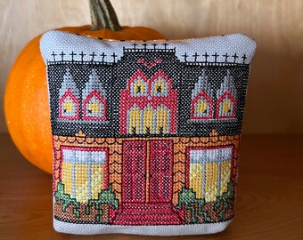 Puede incluir: Una pequeña almohada gris y blanca con un diseño de punto de cruz de una casa encantada con detalles rojos, naranjas y amarillos. La casa tiene tres ventanas y una puerta.