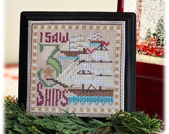 Puede incluir: Un diseño de punto de cruz enmarcado con el texto "I Saw 3 Ships" en hilo rojo. El diseño presenta tres barcos con velas blancas y cascos marrones, navegando en agua azul. El fondo es una tela beige con un borde dorado.