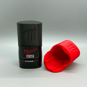 May include: A black Milwaukee M12 REDLITHIUM CP 1.5 battery standing upright next to a bright red protective rubber boot cover. The battery features the Milwaukee logo, M12 branding, and a molded M symbol on the top section of the casing.