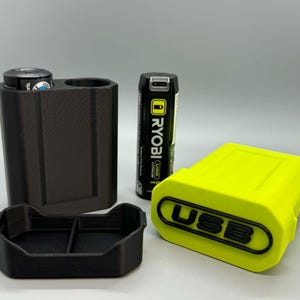 Op de afbeelding: Een zwarte 3D-geprinte batterijhouder met twee sleuven, een bijpassende zwarte basisplaat, een limoengroene batterijafdekking met het woord USB in reli&euml;f in het zwart, en een zwarte Ryobi USB-lithiumbatterij die tussen de houder en de afdekking staat.